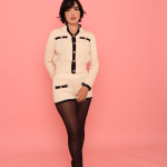 Pessoa usando Conjunto Hani creme com detalhes em preto e botões dourados, composto por blusa de manga longa e shorts combinando, com meia-calça preta e sapatos de salto alto, em frente a fundo rosa.