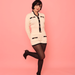 Pessoa usando Conjunto Hani creme com detalhes em preto e botões dourados, composto por blusa de manga longa e shorts combinando, com meia-calça preta e sapatos de salto alto, em frente a fundo rosa.