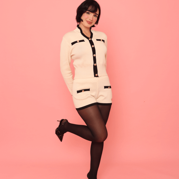 Pessoa usando Conjunto Hani creme com detalhes em preto e botões dourados, composto por blusa de manga longa e shorts combinando, com meia-calça preta e sapatos de salto alto, em frente a fundo rosa.