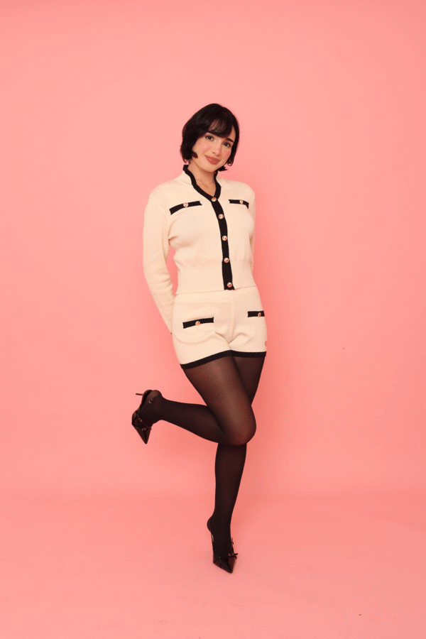 Pessoa usando Conjunto Hani creme com detalhes em preto e botões dourados, composto por blusa de manga longa e shorts combinando, com meia-calça preta e sapatos de salto alto, em frente a fundo rosa.