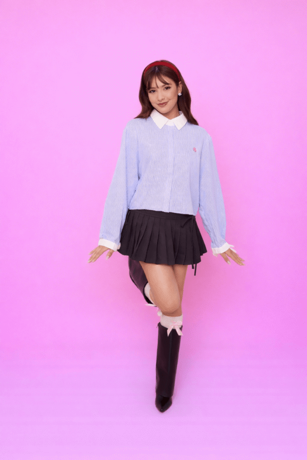 Pessoa usando Saia Yumi preta plissada com detalhes em laço, combinada com camisa listrada azul e branca de manga longa com laço no punho e botas pretas de bico fino, em frente a fundo rosa