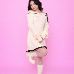 Pessoa usando Vestido Aiko creme ombro a ombro com detalhes em laço preto, meia-calça branca com padrão delicado, polainas brancas e sapatos Mary Jane creme, em frente a fundo rosa.