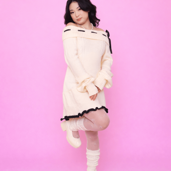 Pessoa usando Vestido Aiko creme ombro a ombro com detalhes em laço preto, meia-calça branca com padrão delicado, polainas brancas e sapatos Mary Jane creme, em frente a fundo rosa.