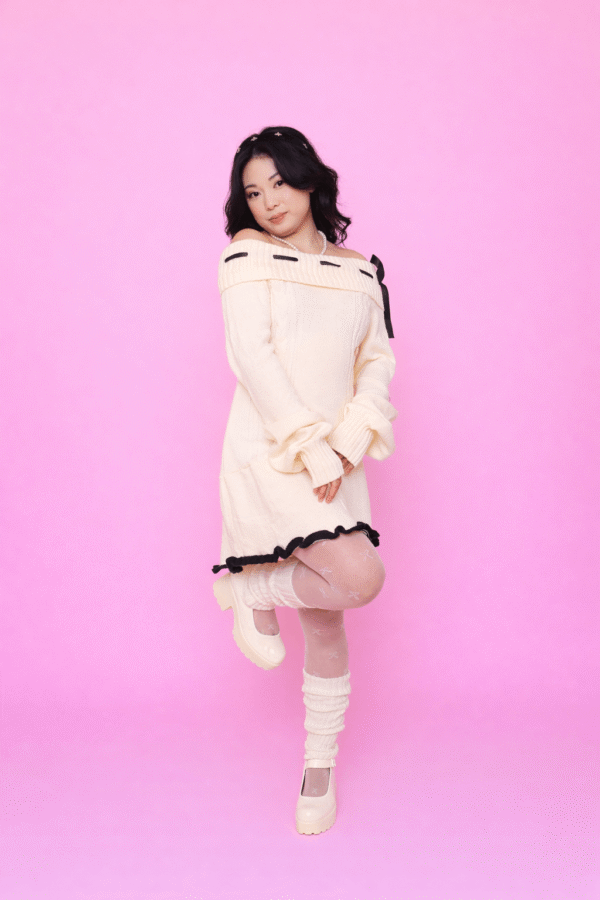 Pessoa usando Vestido Aiko creme ombro a ombro com detalhes em laço preto, meia-calça branca com padrão delicado, polainas brancas e sapatos Mary Jane creme, em frente a fundo rosa.
