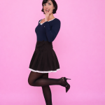 Pessoa usando Blusa Nari azul marinho com renda branca no decote, combinada com saia preta de barra rendada, meia-calça e salto alto, em pose estilosa diante de fundo rosa vibrante.