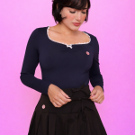 Pessoa usando Blusa Nari azul marinho com renda branca no decote, combinada com saia preta de barra rendada, meia-calça e salto alto, em pose estilosa diante de fundo rosa vibrante.