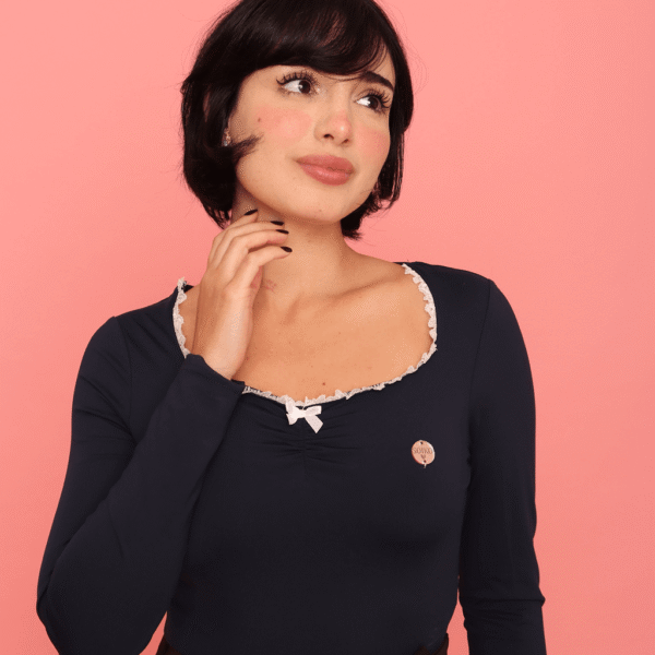 Pessoa usando Blusa Nari azul marinho com renda branca no decote, combinada com saia preta de barra rendada, meia-calça e salto alto, em pose estilosa diante de fundo rosa vibrante.