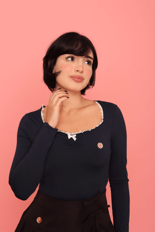 Pessoa usando Blusa Nari azul marinho com renda branca no decote, combinada com saia preta de barra rendada, meia-calça e salto alto, em pose estilosa diante de fundo rosa vibrante.